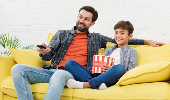 Pai e filho de, aproximadamente, 6 anos, sentados em um sofá amarelo. Eles estão assistindo um filme juntos após um dia com várias tarefas concluídas.