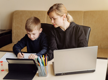 Mãe interessada nos estudos do filho de, aproximadamente, 6 anos, enquanto ele faz o seu dever de casa. Ambos estão sentados em uma mesa branca com folhas, lápis, tablet e computador para pesquisas, em sua superfície.