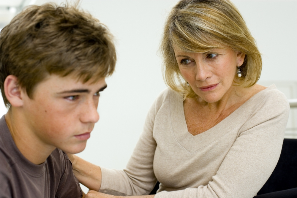 Uma mãe vestindo uma blusa branca acinzentada de manga comprida e conversando com o seu filho sobre as inseguranças dele. Ele tem, aproximadamente, 17 anos e está usando uma camisa marrom.