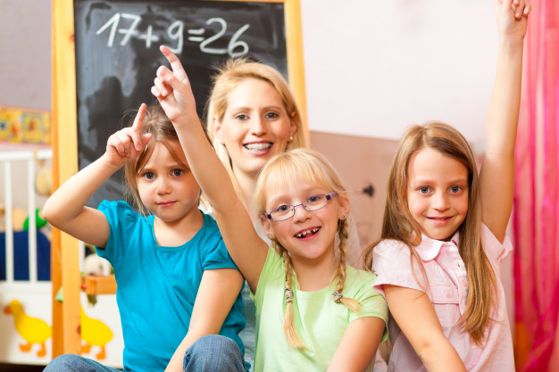 3 meninas de, aproximadamente, 7 anos, (uma de blusa rosa, outra de blusa verde e a terceira de blusa rosa), aprendendo matemática em um quarto colorido e divertido com a presença de uma Tutora.