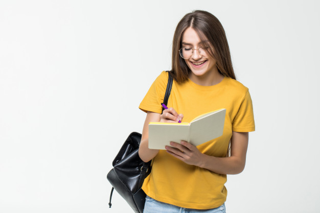 Jovem de, aproximadamente, 17 anos, usando uma blusa amarela, uma calça jeans, um óculos com armação transparente e uma mochila preta. Ela está escrevendo em seu caderno com uma caneta roxa.