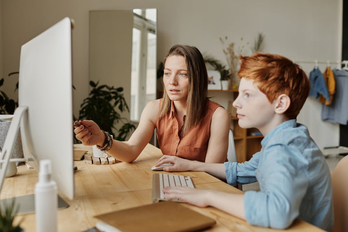 Mãe usando uma blusa sem manga marrom ajudando o seu filho de, aproximadamente, 13 anos, que está usando uma camisa jeans de botão. Eles estão estudando em casa, utilizando um computador que está na superfície de uma mesa de madeira.